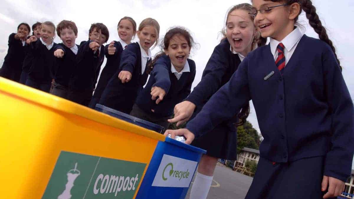 Primary school children in uniform gathered around a compost and recycling bin in a playground