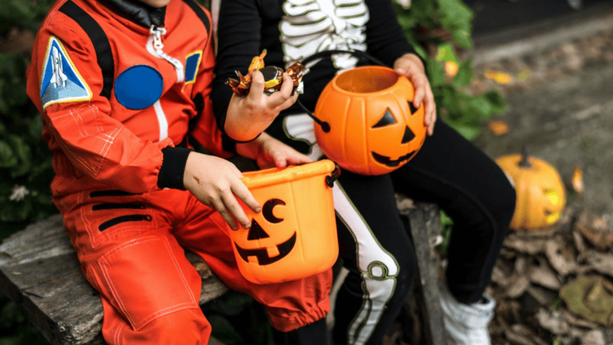 Two children dressed as an astronaut and skeleton sat outside holding plastic pumpkin buckets full of sweets